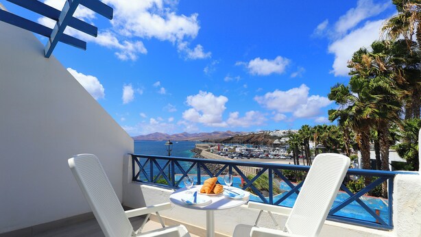 Un balcon avec des chaises blanches et une table, d’où l’on peut admirer une ville côtière sous un ciel bleu dégagé, avec des montagnes à l’horizon.