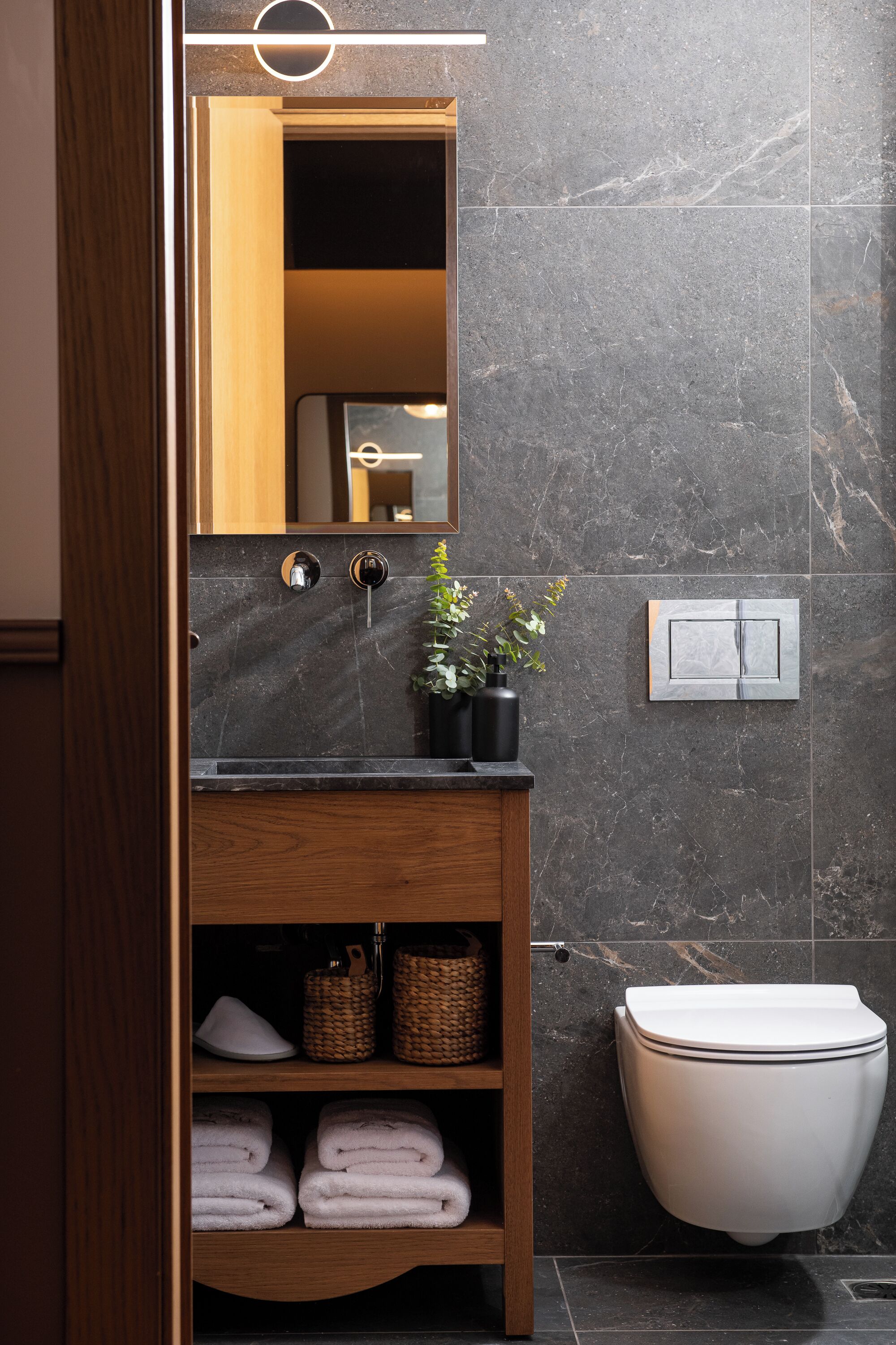 A modern bathroom with a dark marble wall, a wooden vanity with a mirror, a sink, and a toilet. There are baskets and folded towels on the vanity.