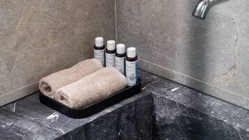 Un baño con lavamanos de mármol, un perchero con toallas y botellas, y un cabezal de ducha.