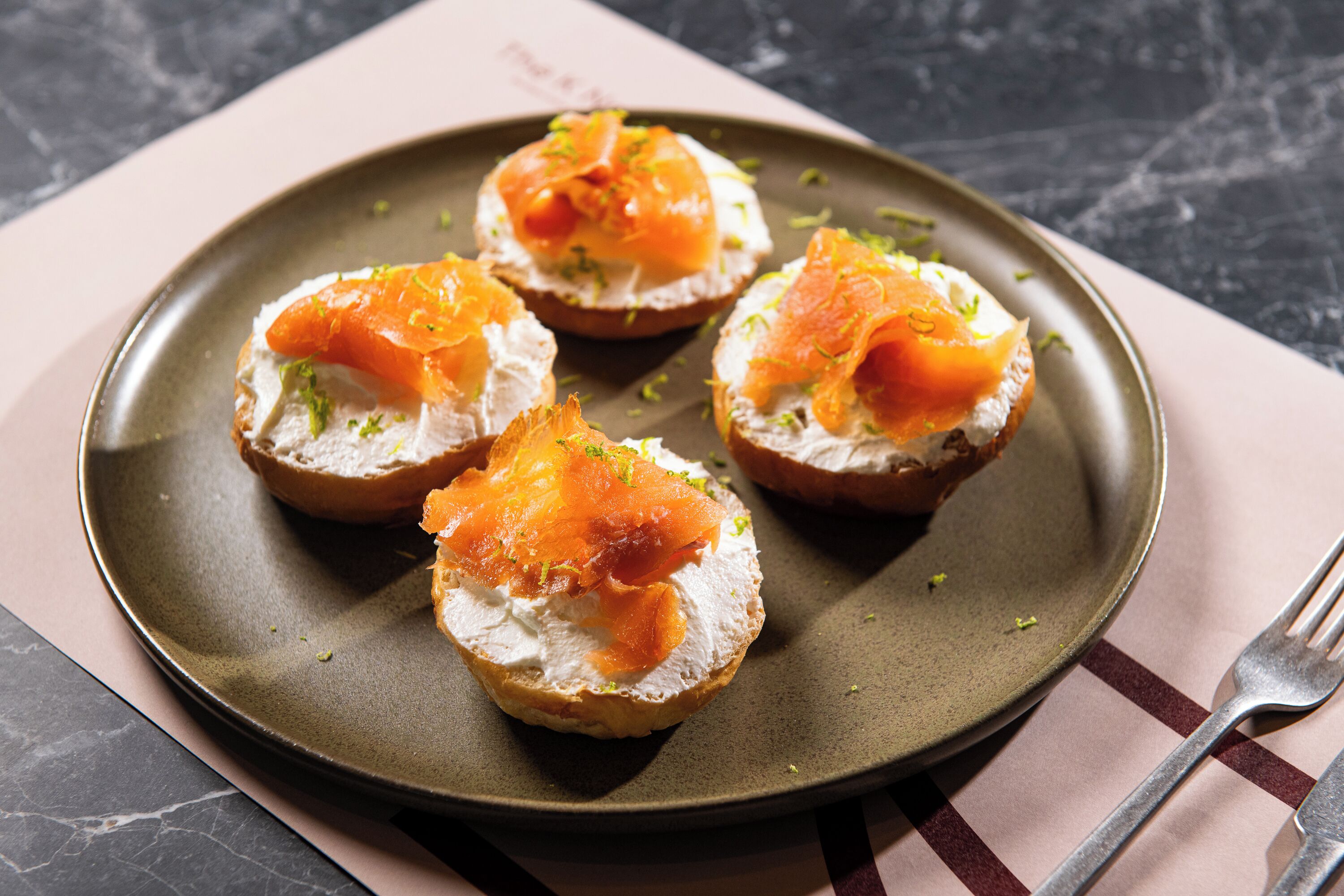 A plate with four open-faced sandwiches topped with smoked salmon and garnished with herbs, placed on a dark plate with a fork beside it.
