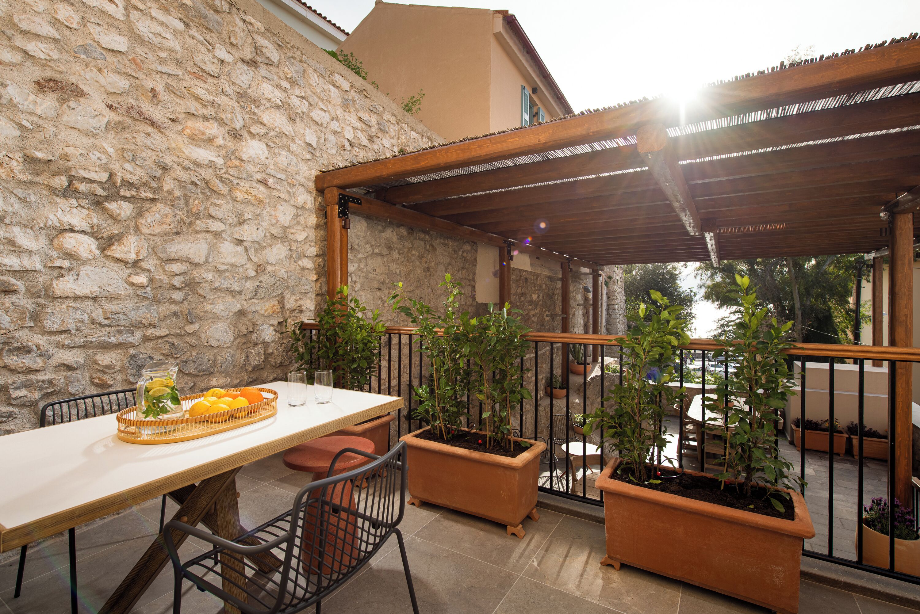 Un muro de piedra, una pérgola de madera, una mesa al aire libre, sillas, plantas en macetas y vista al mar.