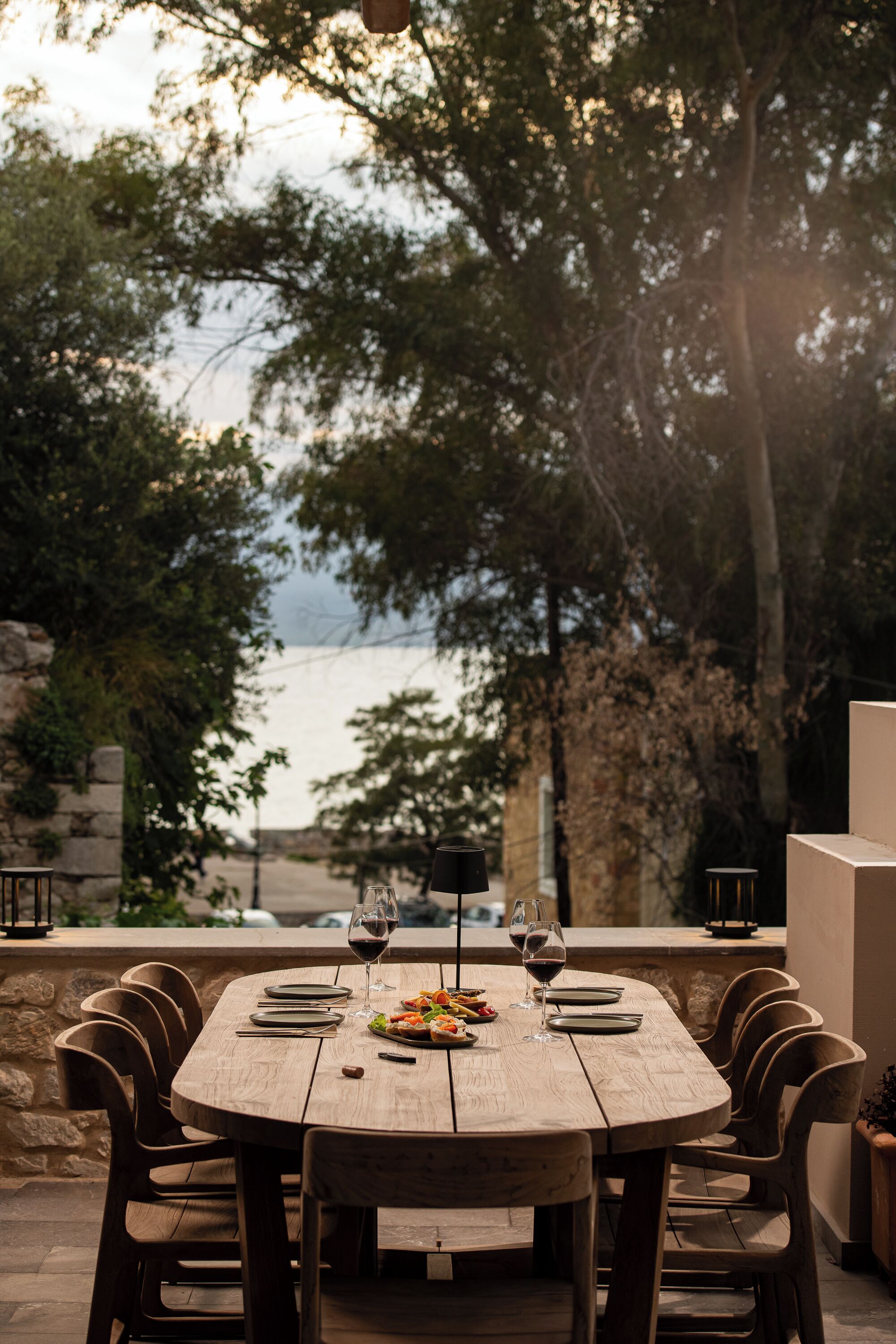 An outdoor dining area with a wooden table and chairs, set against a backdrop of trees and a view of the sea.