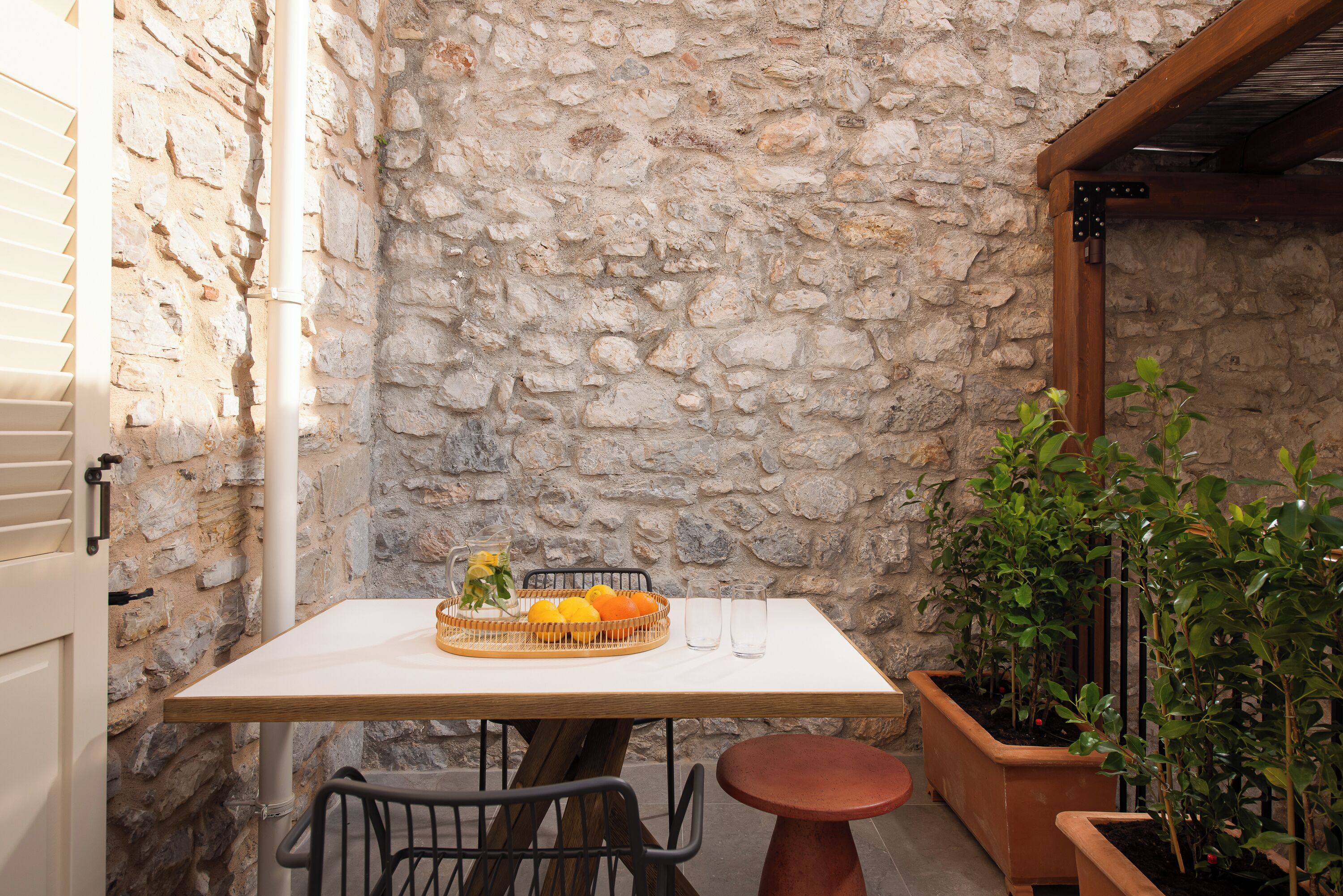 Un muro de piedra, una mesa con fruteros, dos sillas, una pequeña mesa redonda y plantas en macetas.