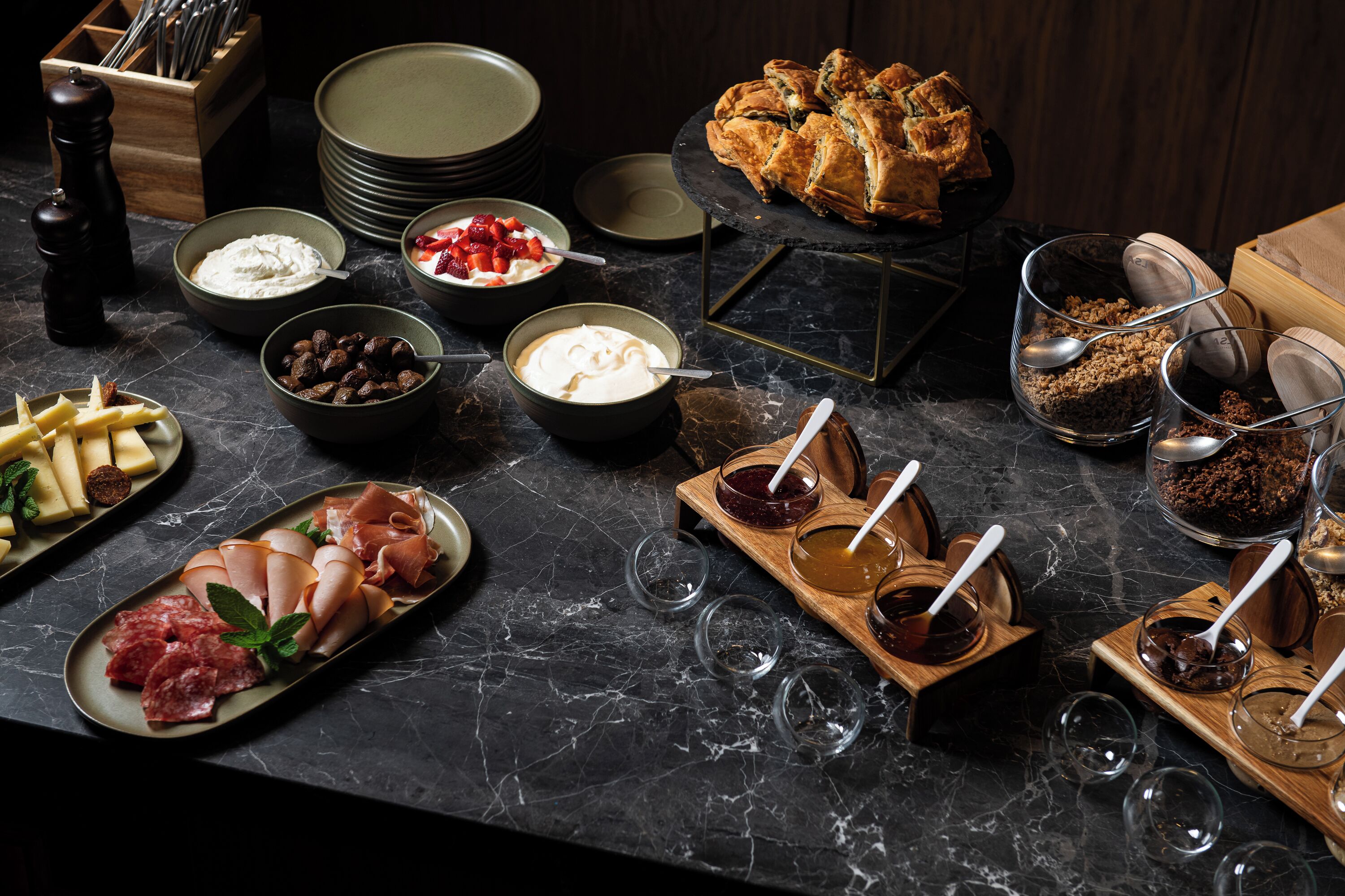 A variety of food items and utensils on a marble countertop, including bowls of cream and berries, a plate of sliced meats, and a loaf of bread.