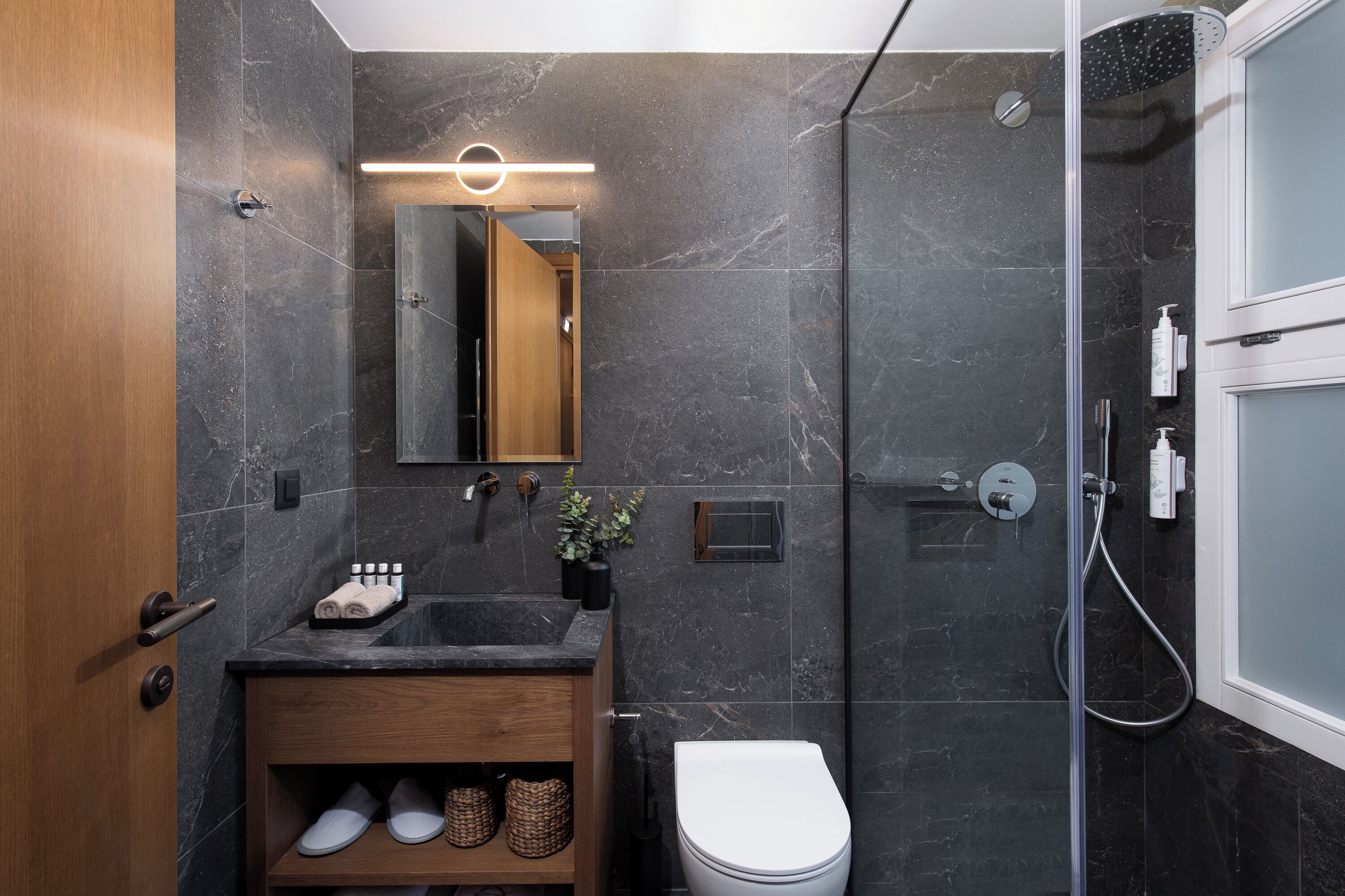 A modern bathroom with a marble wall, a wooden vanity, a white toilet, and a glass shower enclosure.