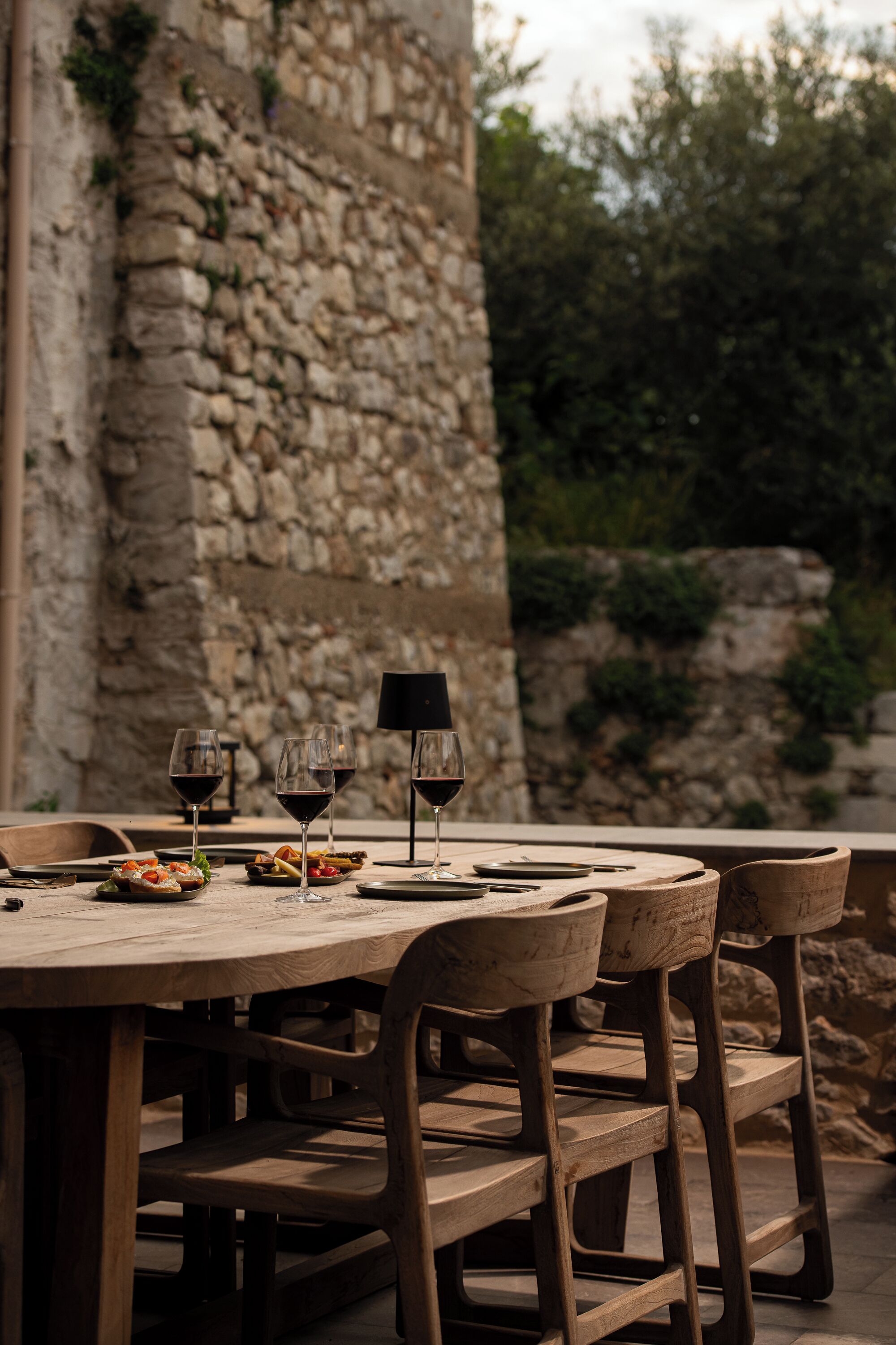 A rustic outdoor dining table with wooden chairs, set against a stone wall backdrop.