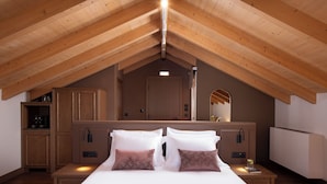 A bedroom with a sloped wooden ceiling, a large bed with white linens and two decorative pillows, a nightstand with a lamp, and a wooden wardrobe.