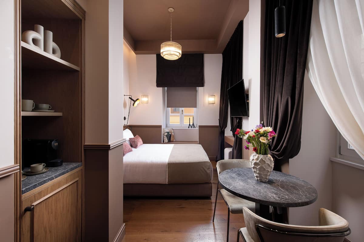 Ett kompakt hotellrum med en säng, ett litet matbord med stolar, en kaffemaskin och en vas med blommor.