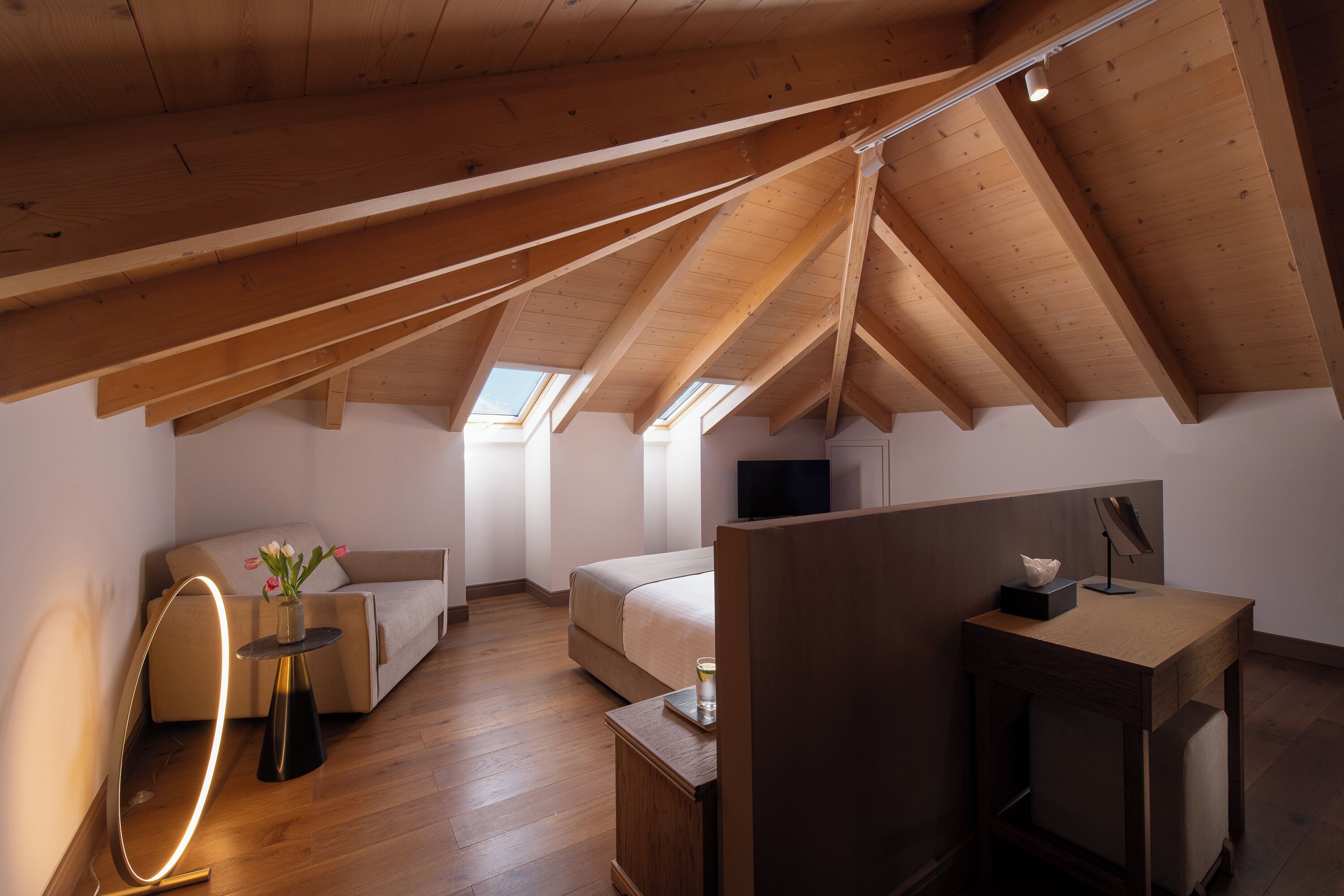 Un dormitorio tipo loft moderno con techo inclinado, tragaluces, cama, escritorio y una pequeña mesa redonda con lámpara.