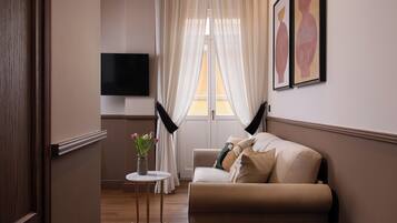 A modern living room with a beige sofa, a small round table with a vase of flowers, and a flat-screen TV mounted on the wall.