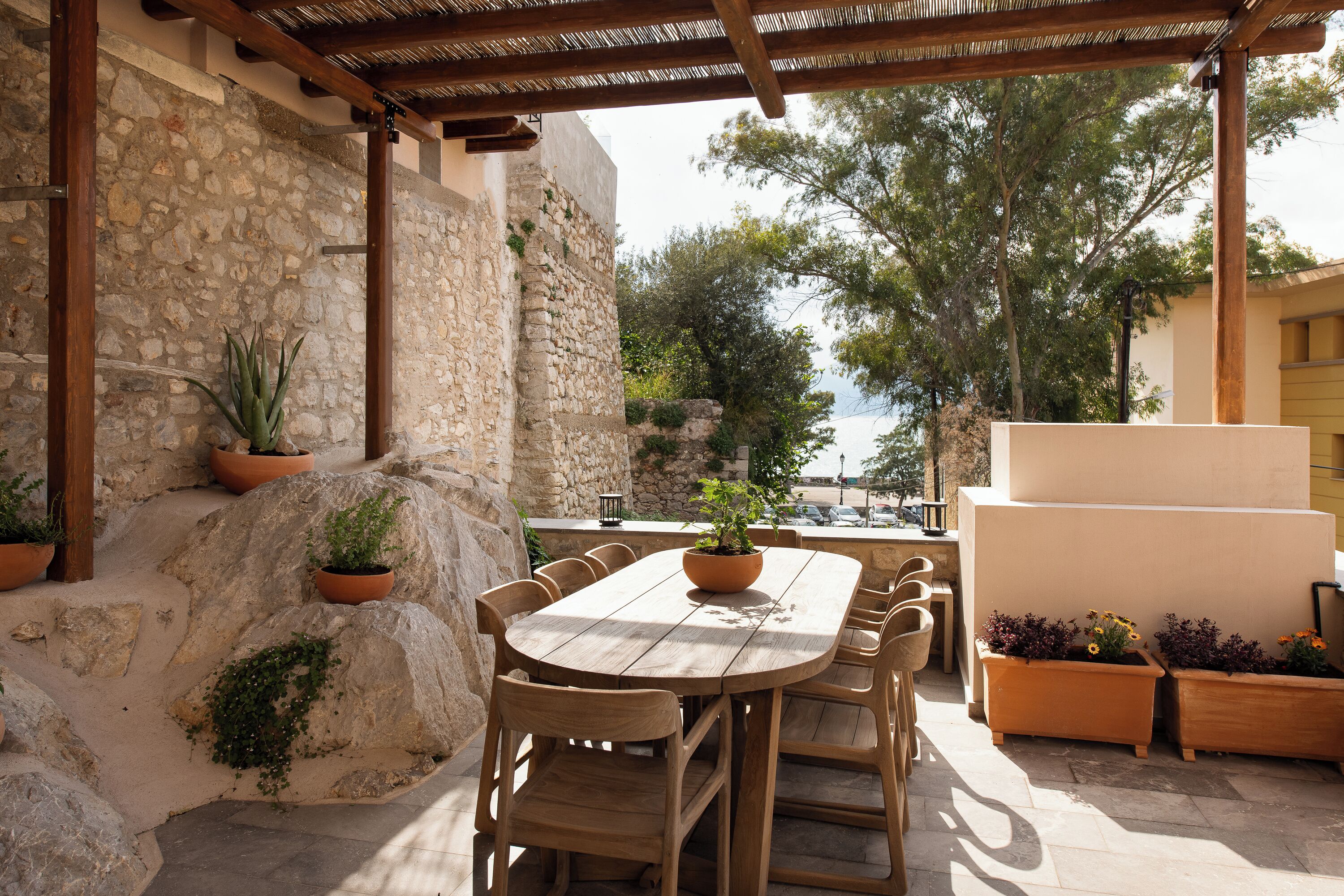 An outdoor patio with a stone wall, wooden table, and chairs, surrounded by potted plants and overlooking a scenic view.