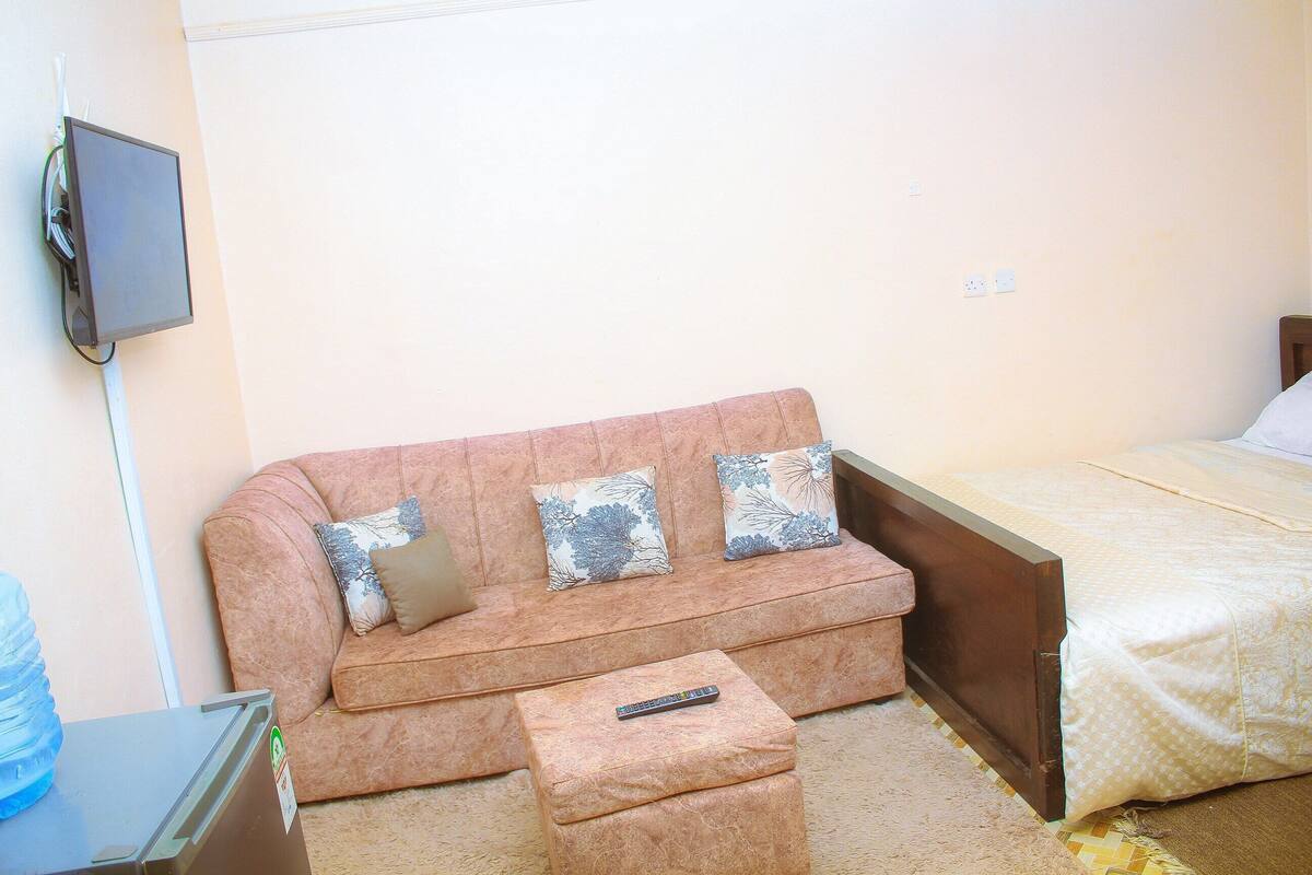 A room with a sofa, a bed, a remote control on a small table, and a TV mounted on the wall.