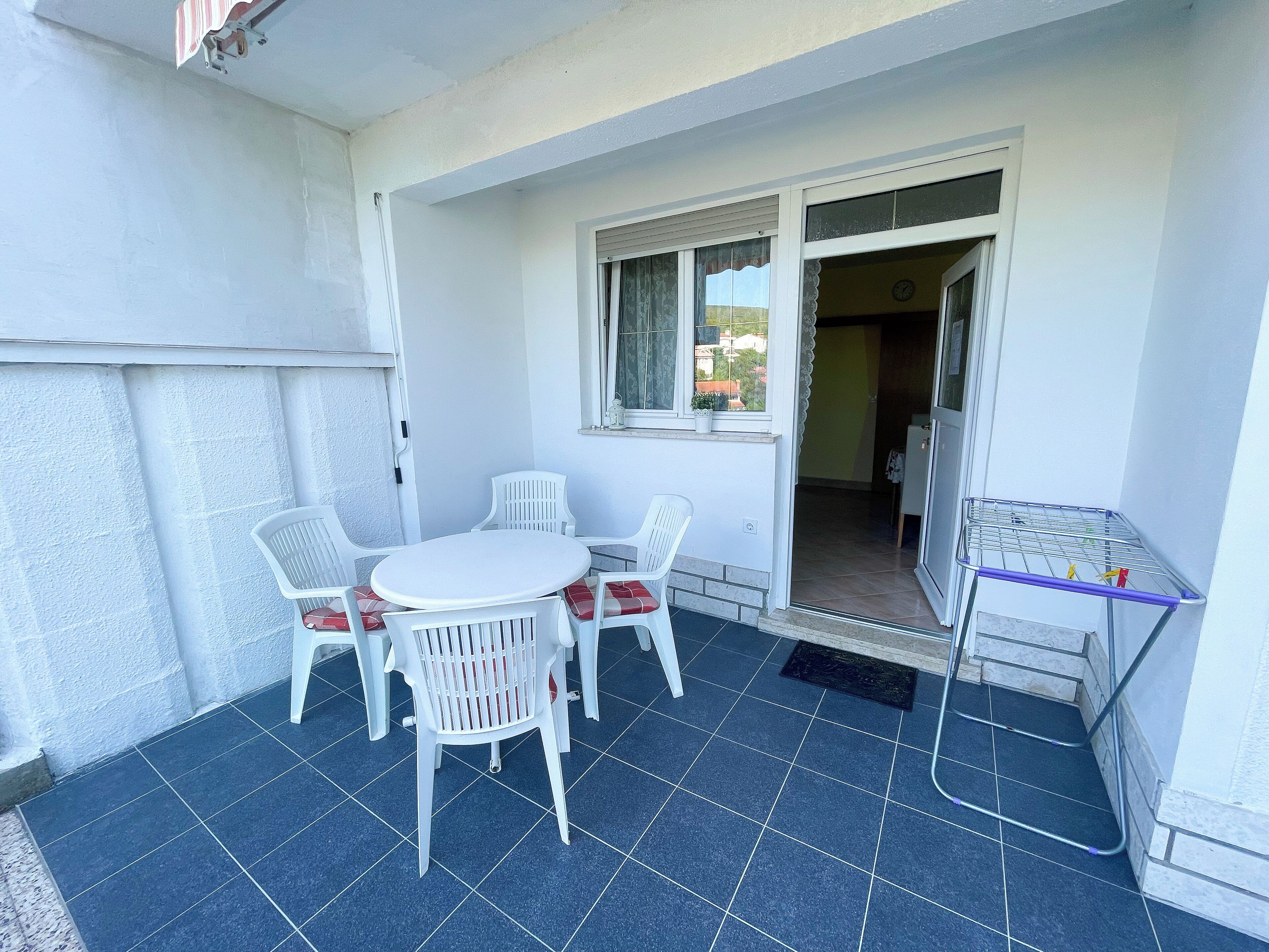 A small patio area with a round table and four chairs, a window with curtains, and a metal drying rack.