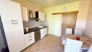 A compact kitchen with white cabinets, a black countertop, and a dining area with a table and chairs. - Apartment Combaj 3 (Jadranovo)