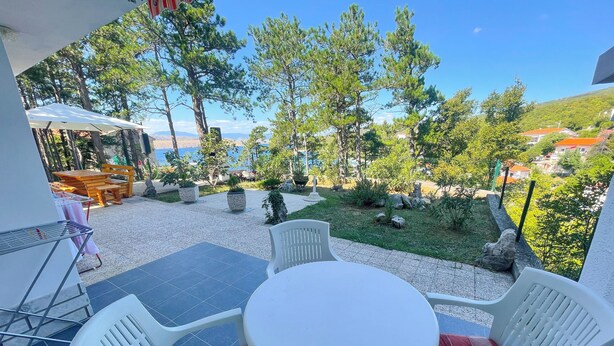 A patio with a table and chairs, a view of the sea, and a garden with trees and plants. - Apartment Combaj 3 (Jadranovo)