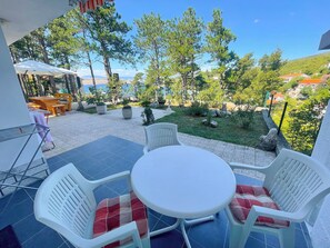 A patio with a table and chairs, a view of the sea, and a garden with trees and plants. - Apartment Combaj 3 (Jadranovo)