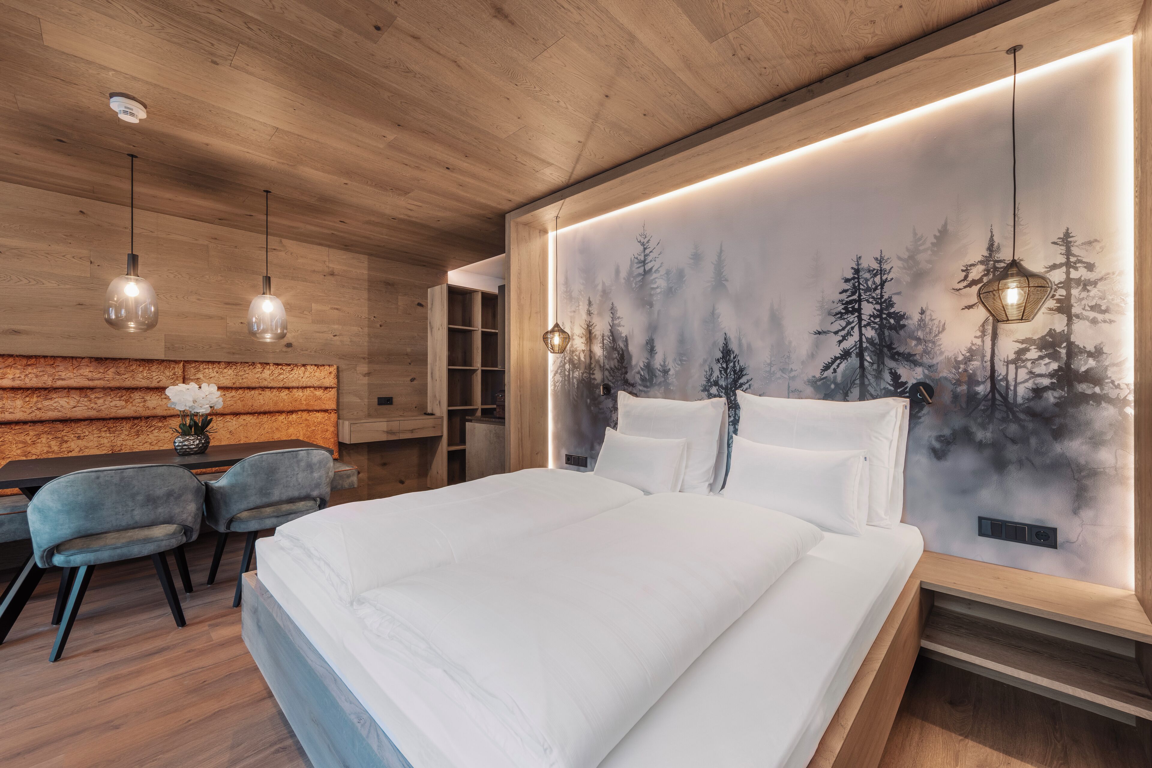 A modern bedroom with a wooden ceiling, a large bed, a desk with chairs, and a wall mural of a forest.