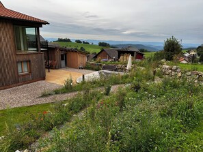 Una casa de madera rústica con un balcón, una pared de piedra y un jardín lleno de plantas y flores diversas.