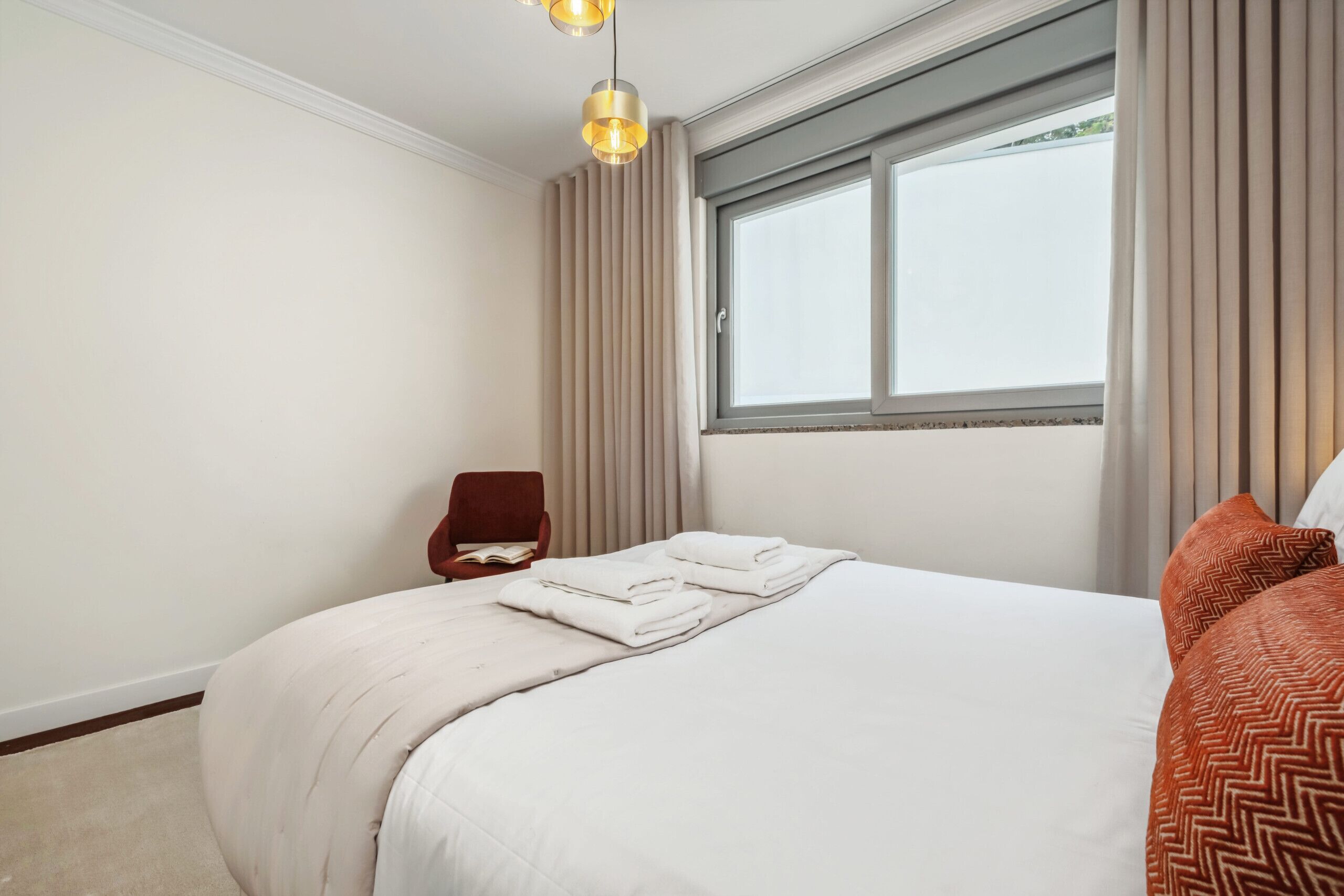 A hotel room with a neatly made bed, a window with curtains, a red chair, and a ceiling light fixture.