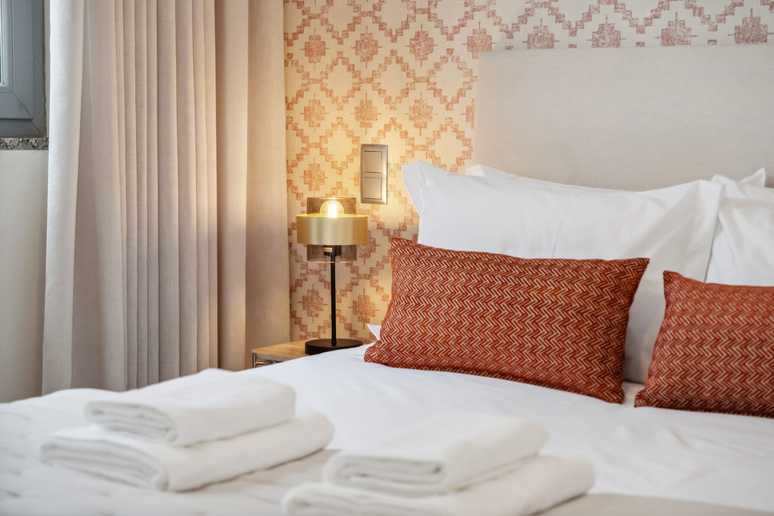 A neatly made bed with white linens, a lamp on the nightstand, and patterned wallpaper.