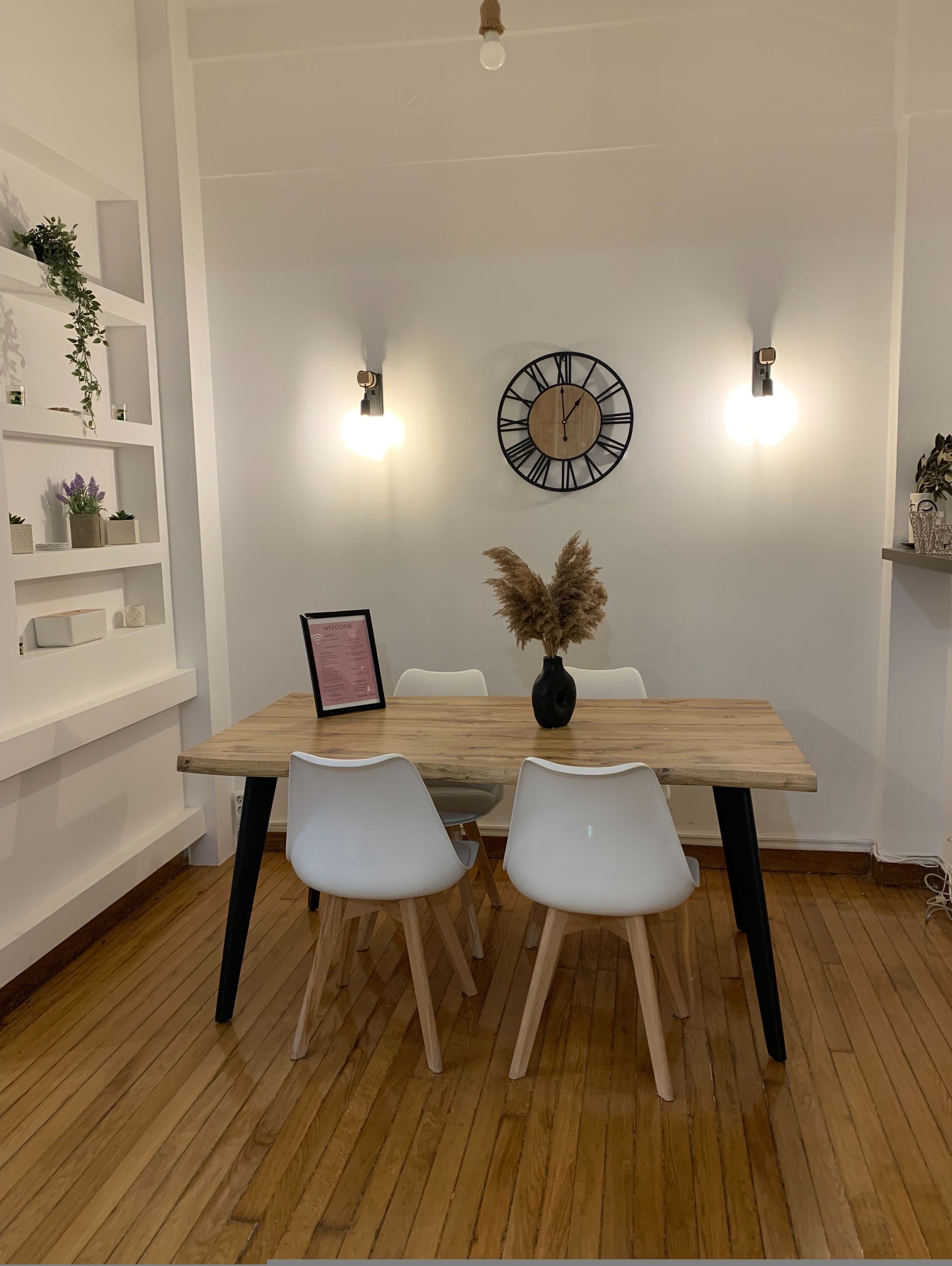 Uma sala de jantar com mesa de madeira, cadeiras brancas, um relógio e luminárias na parede.