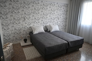 Een slaapkamer met een bed, twee kussens, een nachtkastje, een lamp, een behang met patroon, een raam met gordijnen, en een gevlekte vloer.