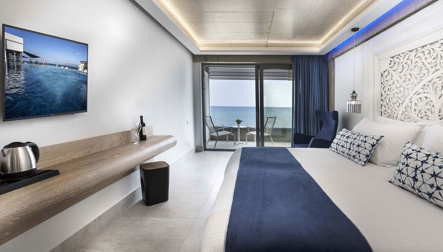 A modern hotel room with a large bed, a seating area with a view of the sea, a flat-screen TV, and a balcony with a glass door.
