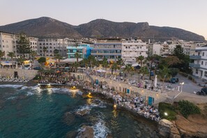 Hotel frente al mar con terraza exterior, una pared de piedra y una montaña al fondo.