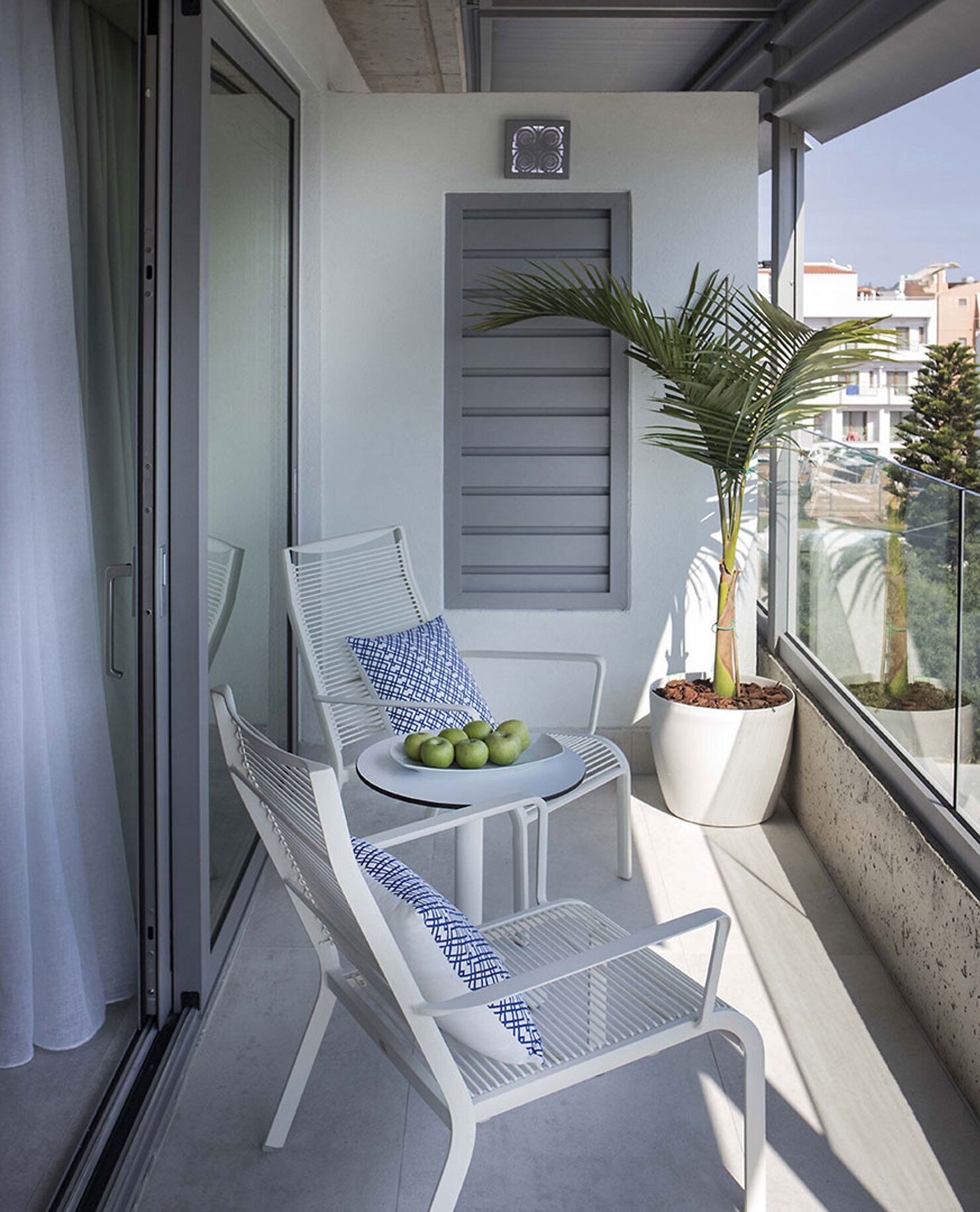 A balcony with a white metal chair, a small round table with a bowl of green apples, and a potted palm plant.