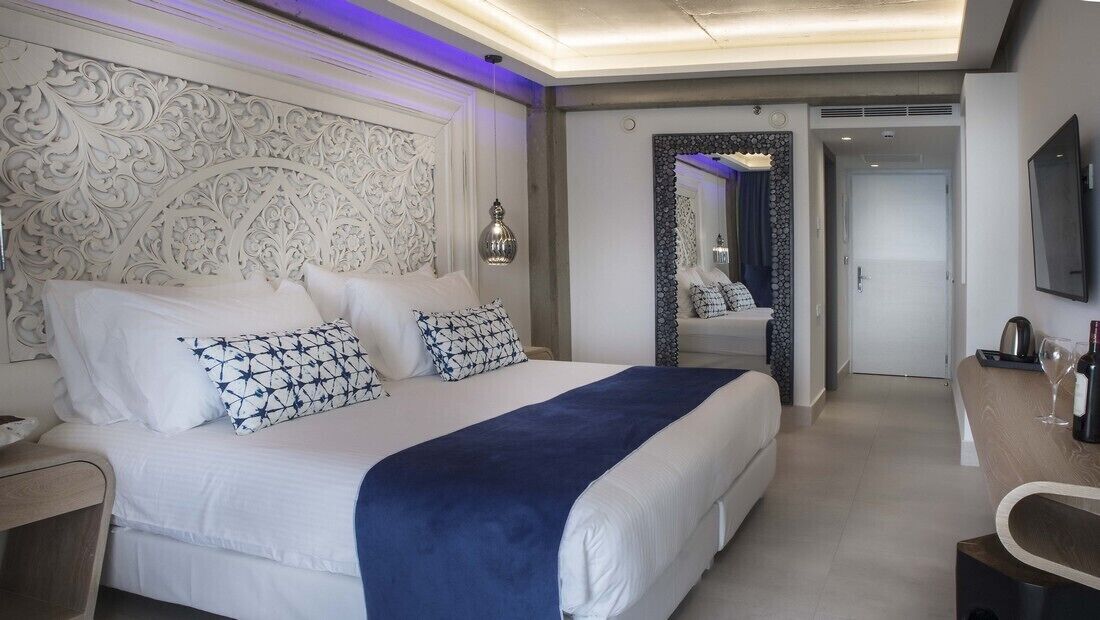 Quarto de hotel com uma cama grande, uma escrivaninha, um espelho e uma televisão.