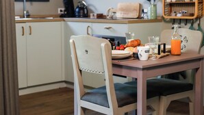 A dining area with a wooden table, two chairs, a television mounted above the table, a kitchen area with cabinets and a sink, and a window providing natural light. - Romantic luxury apartment  great garden with fire table 5 minutes to the forest. (Wilbertoord)