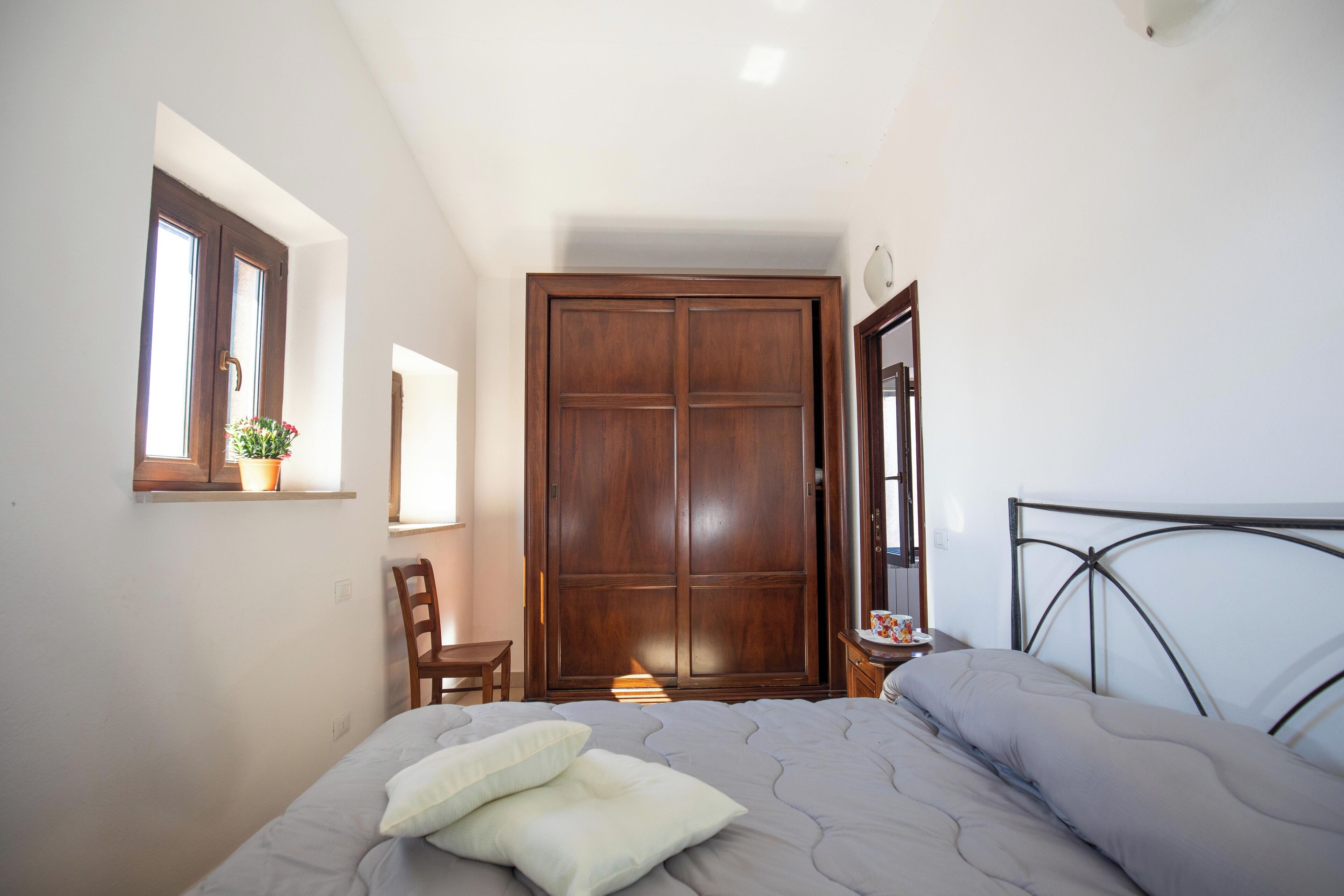 A bedroom with a bed, a wooden wardrobe, a chair, a small table, and a window with a plant.