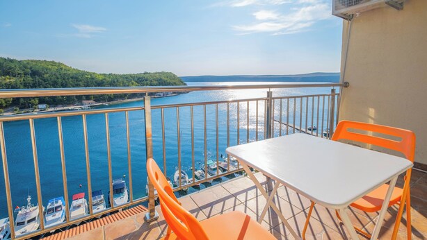 A balcony with a white table and orange chairs, overlooking a marina with boats and a distant island. - Apartment Ahel (Jadranovo)