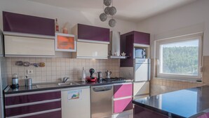 A modern kitchen with purple and white cabinetry, stainless steel appliances, and a window with a forest view. - Apartment Ahel (Jadranovo)