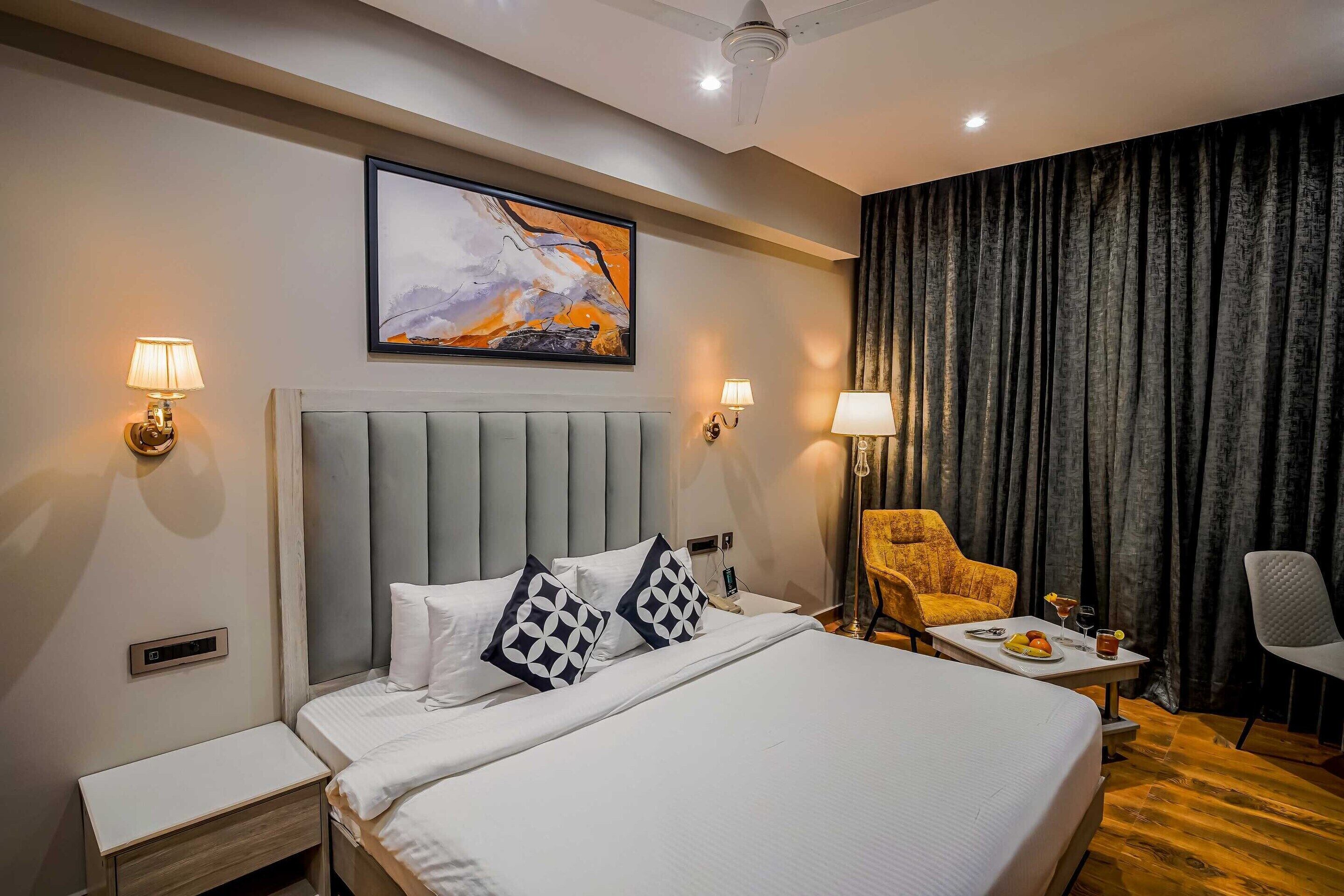 Une chambre d’hôtel avec un grand lit, deux lampes de chevet, une tête de lit, un tableau au mur, une chaise, une petite table avec une assiette de fruits et une boisson, et un rideau.