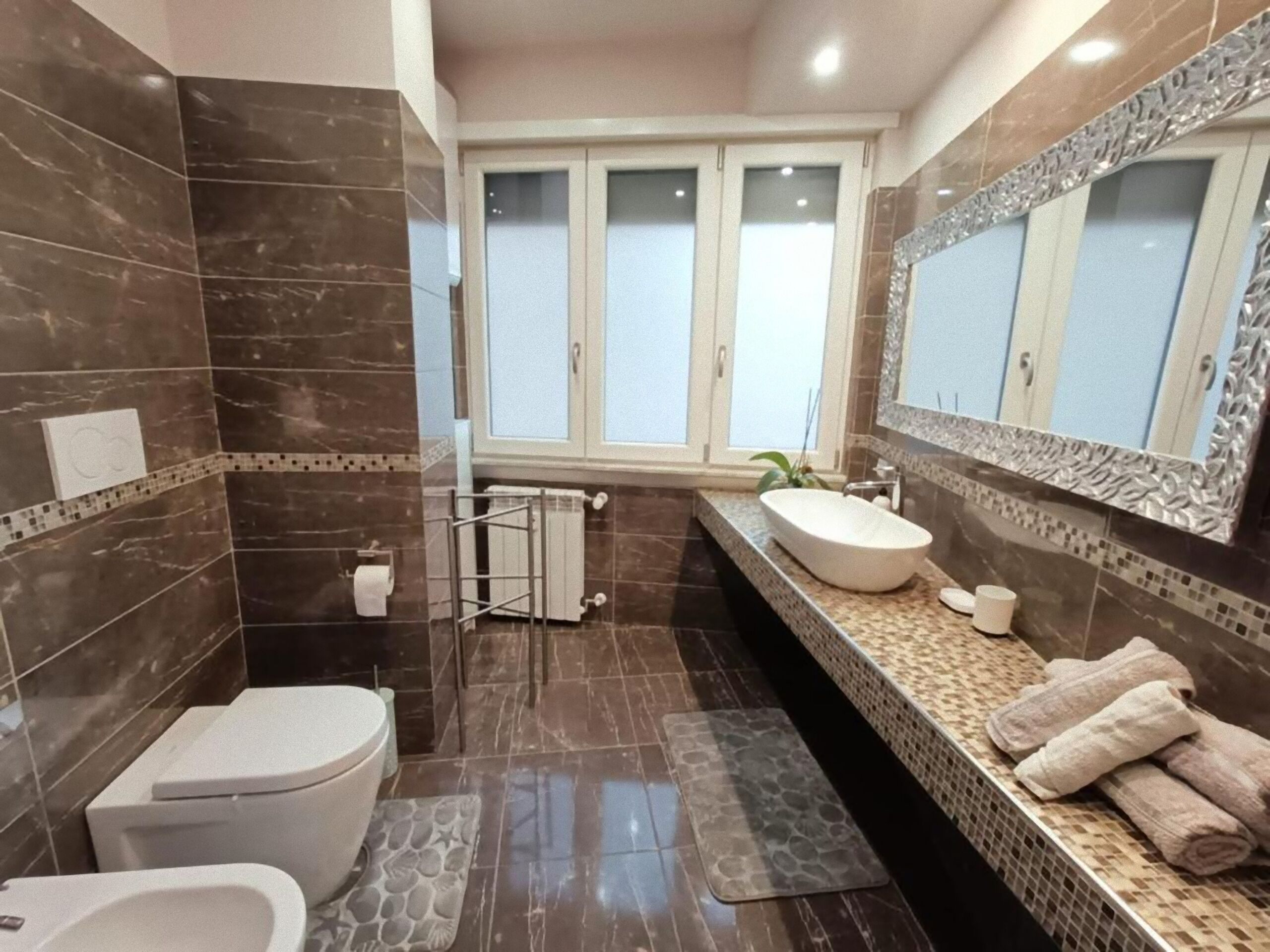 Casa de banho moderna com azulejos de mármore escuro, sanita branca, lavatório com espelho grande e janela com portas de vidro jateado.