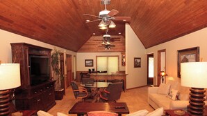 Uma sala de estar espaçosa com teto alto, detalhe em madeira no teto, uma TV grande, um espaço de jantar com mesa e cadeiras, e um sofá com mesinha de centro.