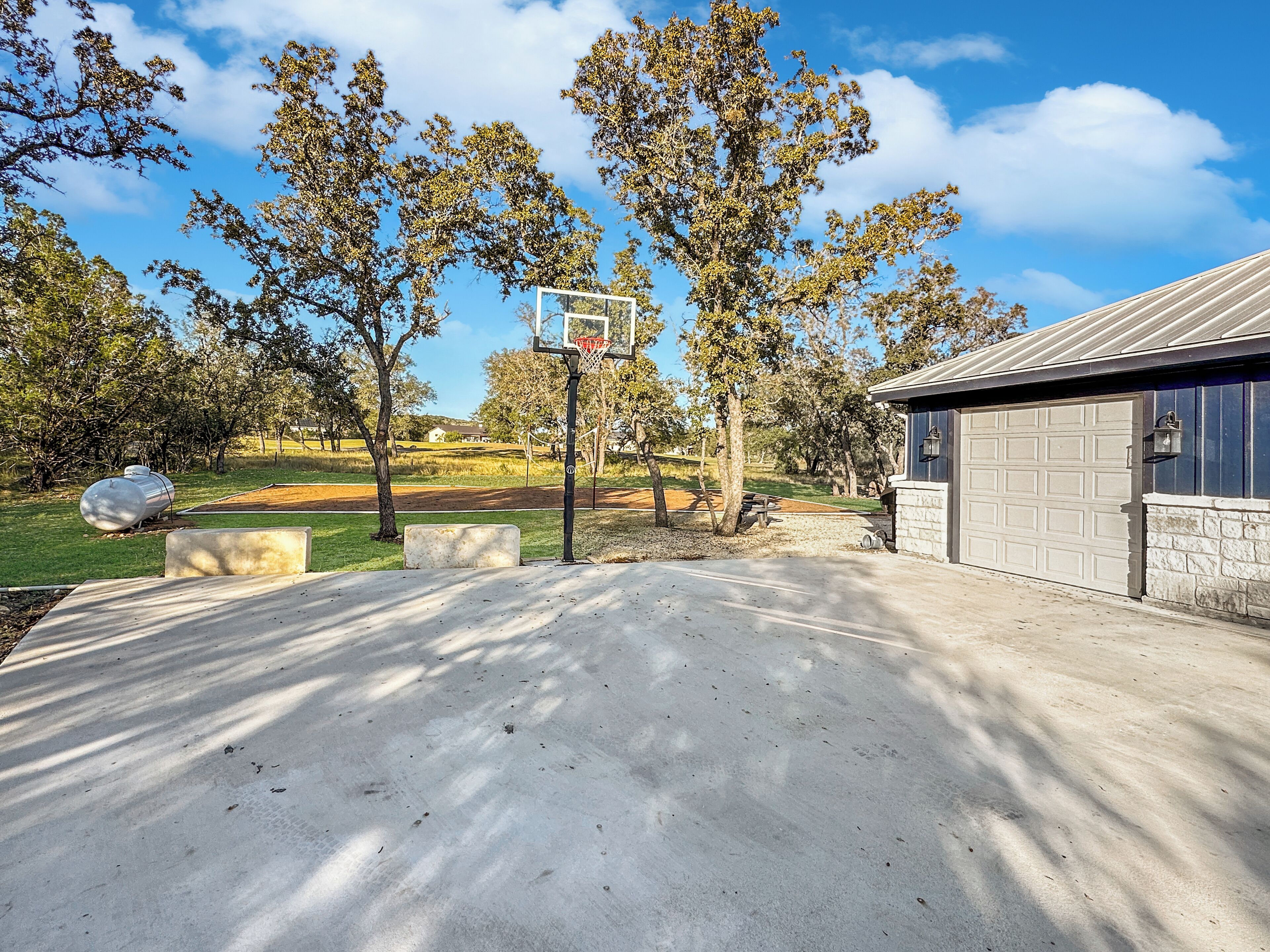 A basketball court with a hoop, trees, and a garage.