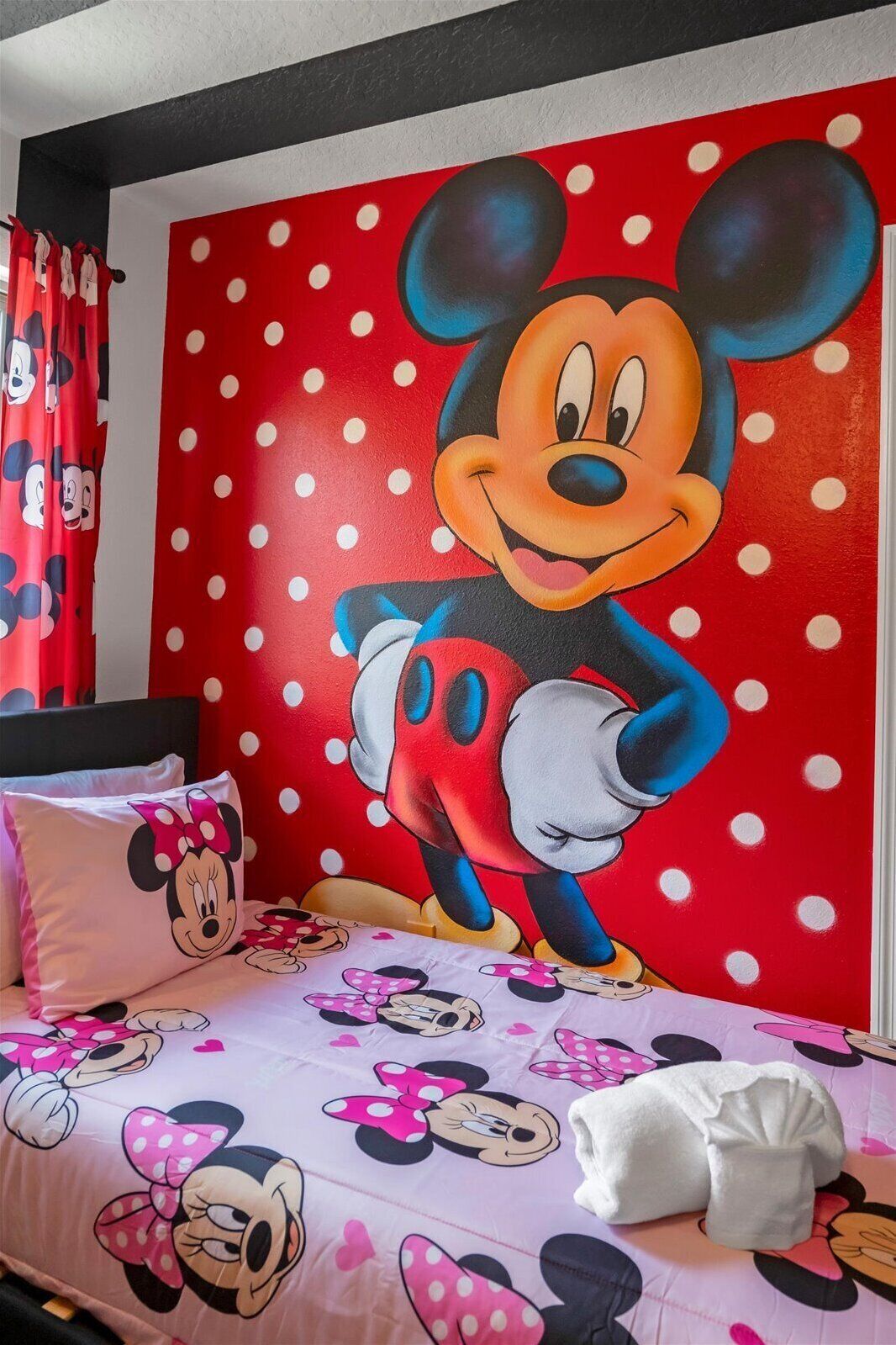 A bedroom with a large Mickey Mouse mural on the wall, a bed with Minnie Mouse bedding, and red curtains with Mickey Mouse prints.