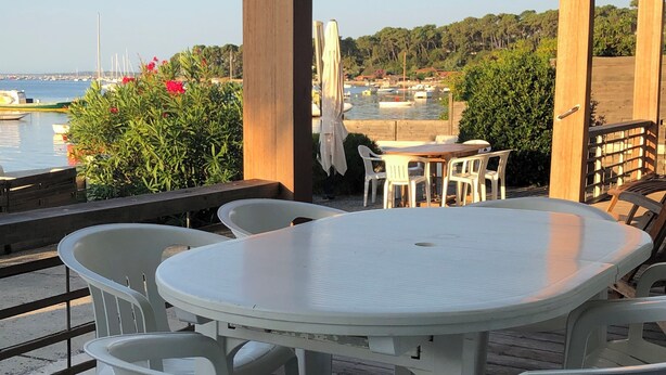 An outdoor patio with a round table and white plastic chairs, overlooking a marina with boats and greenery.