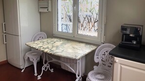 A compact kitchen with a refrigerator, a window with decorative blinds, a marble table, two white chairs, and a coffee machine.