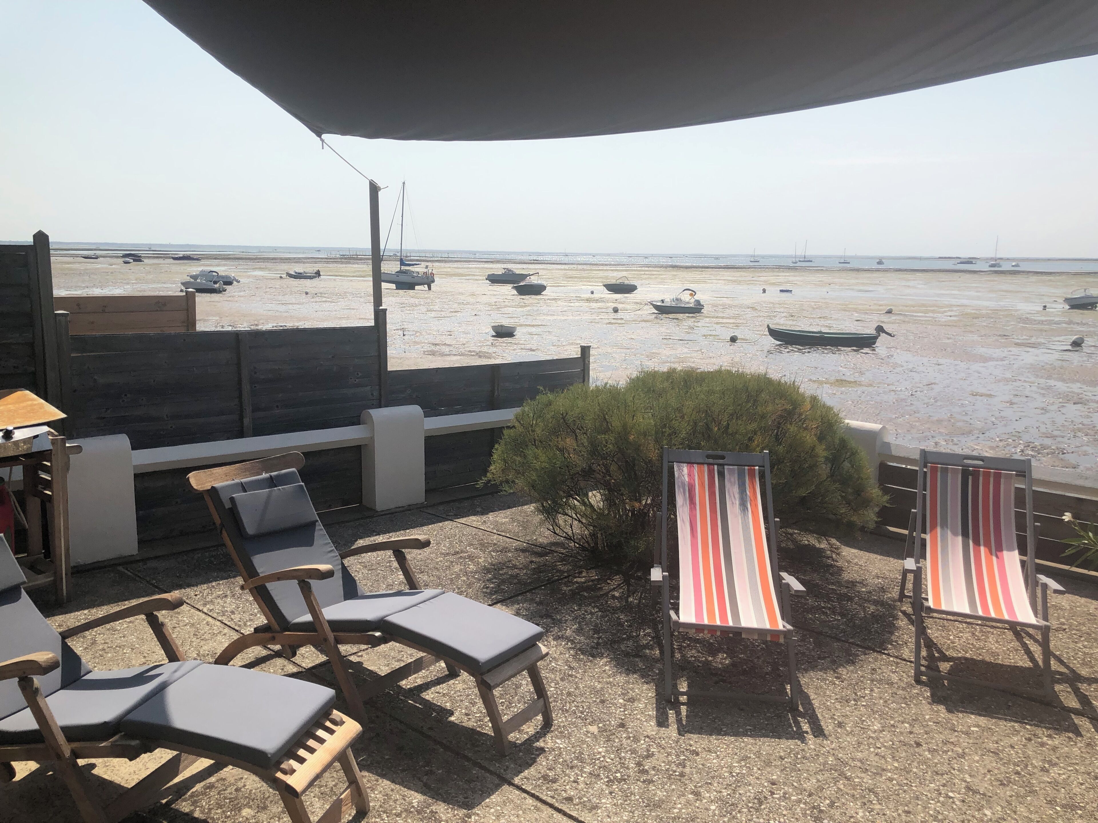 En uteplats vid stranden med solstolar, ett litet bord och utsikt över båtar på vattnet.