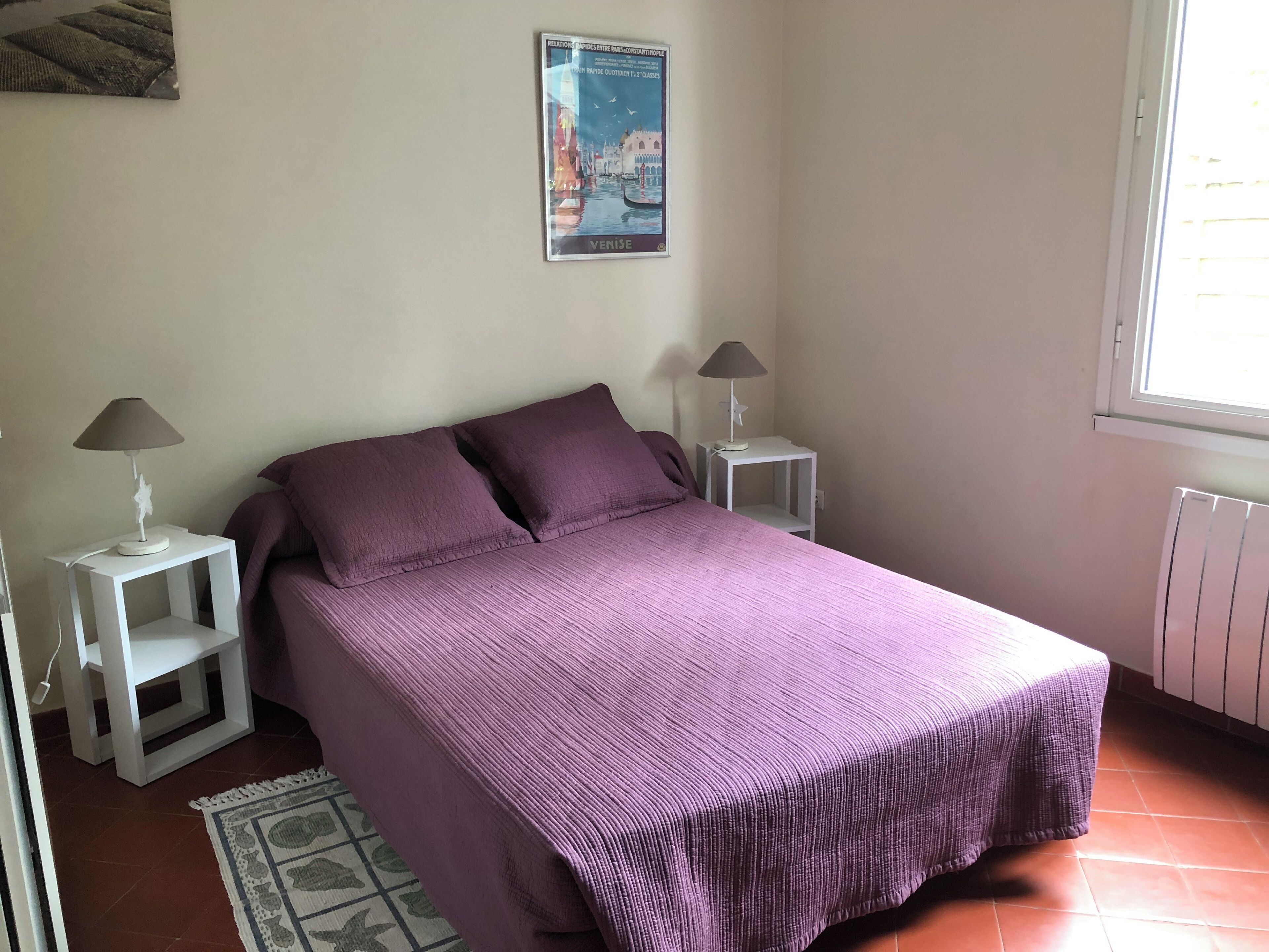 A bedroom with a large bed, two bedside tables with lamps, a poster on the wall, and a window with blinds.