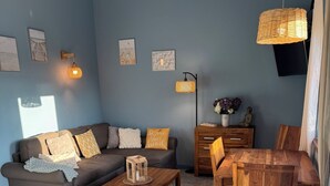 A living room with a sofa, coffee table, wooden side table, and a dining table with chairs. There are wall-mounted lamps and framed pictures on the wall.