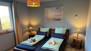 A double bed with two pillows and a blanket, a bedside table with a lamp, a painting on the wall, and a window with curtains.