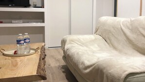 Een kamer met een witte bank, een houten bijzettafeltje met waterflesjes, een televisie op een plank en een witte kledingkast.