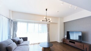 A modern living room with a sofa, a round coffee table, a TV on a wooden stand, and a large window with curtains. - Creare Togoshi Ginza by Tranova (Tokyo)