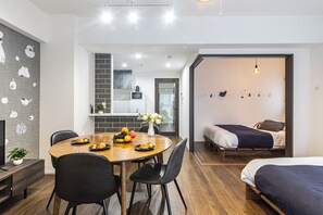 A modern, well-lit room with a wooden dining table, black chairs, a bed with a dark blue blanket, and a small kitchenette in the background. - Creare Togoshi Ginza by Tranova (Tokyo)