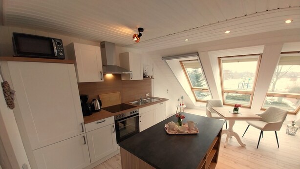 A modern kitchen with a central island, white cabinetry, and a dining area with a table and chairs. - Vacation apartment "Still Hörn 3" in Greetsiel (approx. 35 sqm / 2 persons) (Krummhörn)