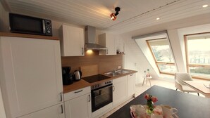 A modern kitchen with white cabinets, a wooden countertop, a stove, an oven, and a microwave. There's a dining area with a table and chairs, and a window with a view. - Vacation apartment "Still Hörn 3" in Greetsiel (approx. 35 sqm / 2 persons) (Krummhörn)